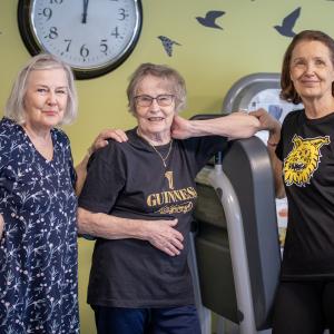 Kolme seniori-ikäistä hymyilevää rouvaa sseisoo vierekkäin kuntosalilla.